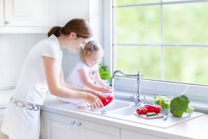 Young beautiful mother and her cute curly toddler daughter washing vegetables together in a kitchen sink getting ready to cook salad for lunch in a sunny white kitchen with a big garden view window