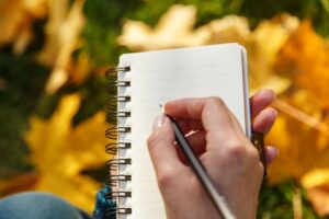 A woman's hand holding a paper notebook and pencil against the yellow leaves in the soft sunlight. Autumn mood, empty space for text