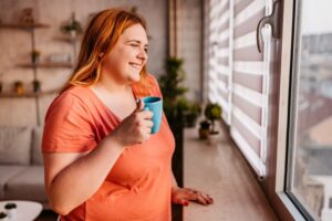 plus-size-woman-looks-out-window-holding-coffee-mug